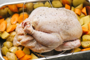 A whole trussed chicken on a trivet in a roasting pan surrounded by chopped carrots and potatoes.