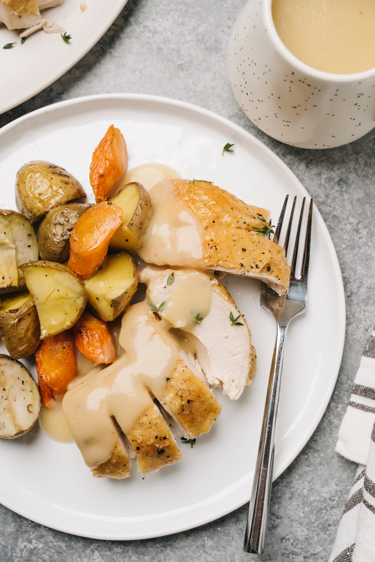 Sliced roasted chicken breast on a plate drizzled with gravy, with roasted potatoes and carrots and a small pitcher of gravy to the side.