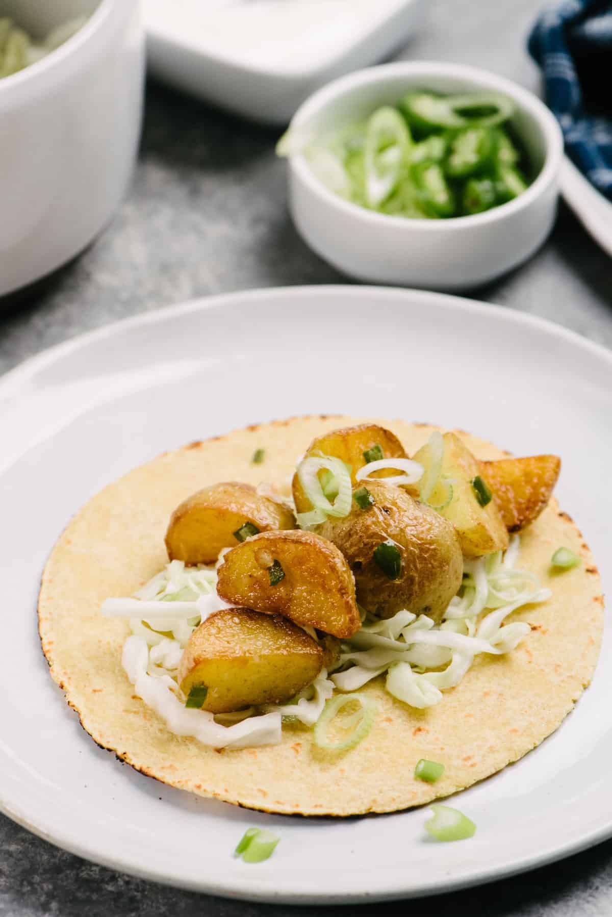 Side view, a spicy potato taco on a plate garnished with coleslaw and serrano chilies; sliced green onions in a bowl and a blue napkin are in the background.