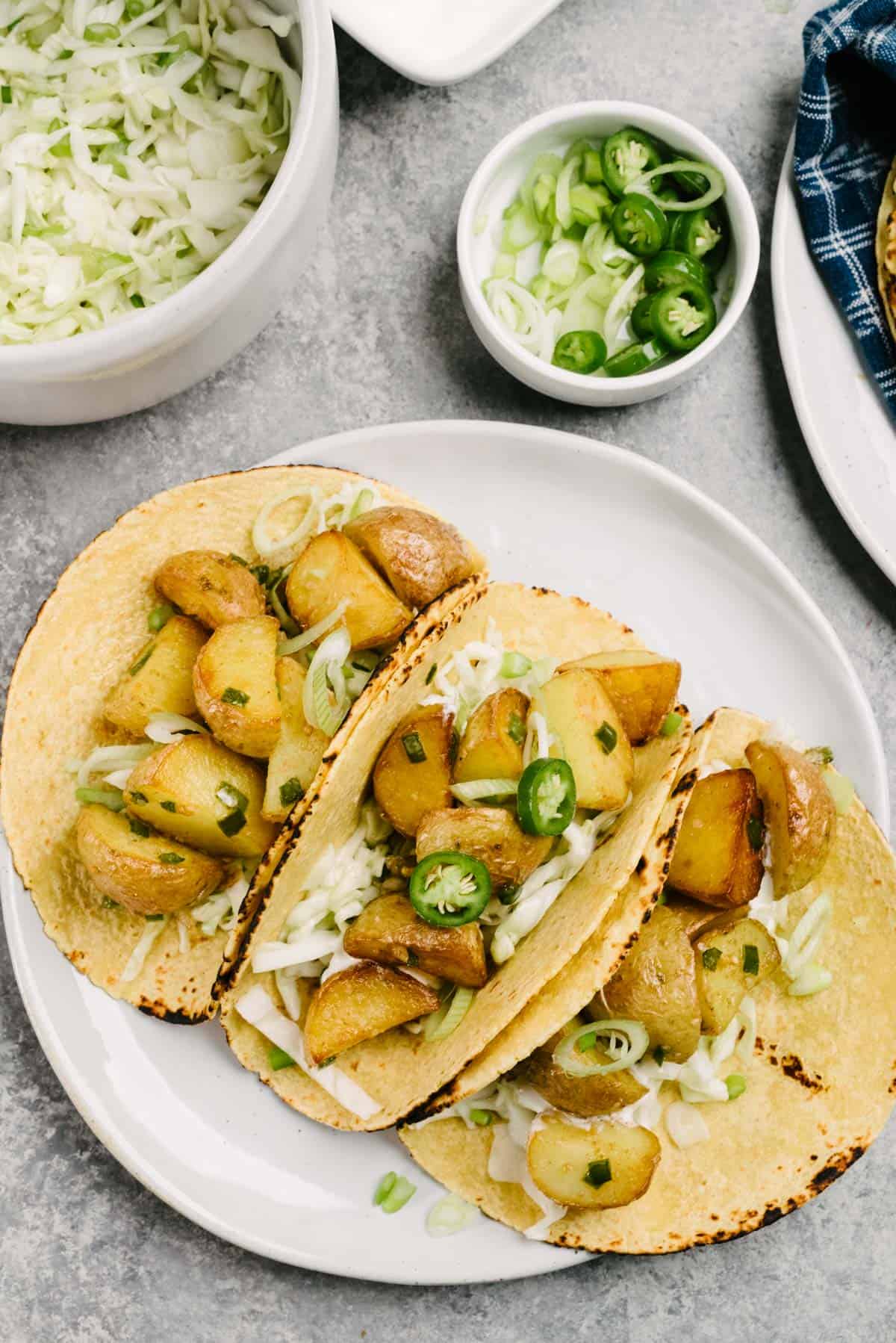 Three potato tacos in corn tortilla shells on a plate, garnished with coleslaw, serrano chilies, and green onions; small bowls of toppings surround the plate.