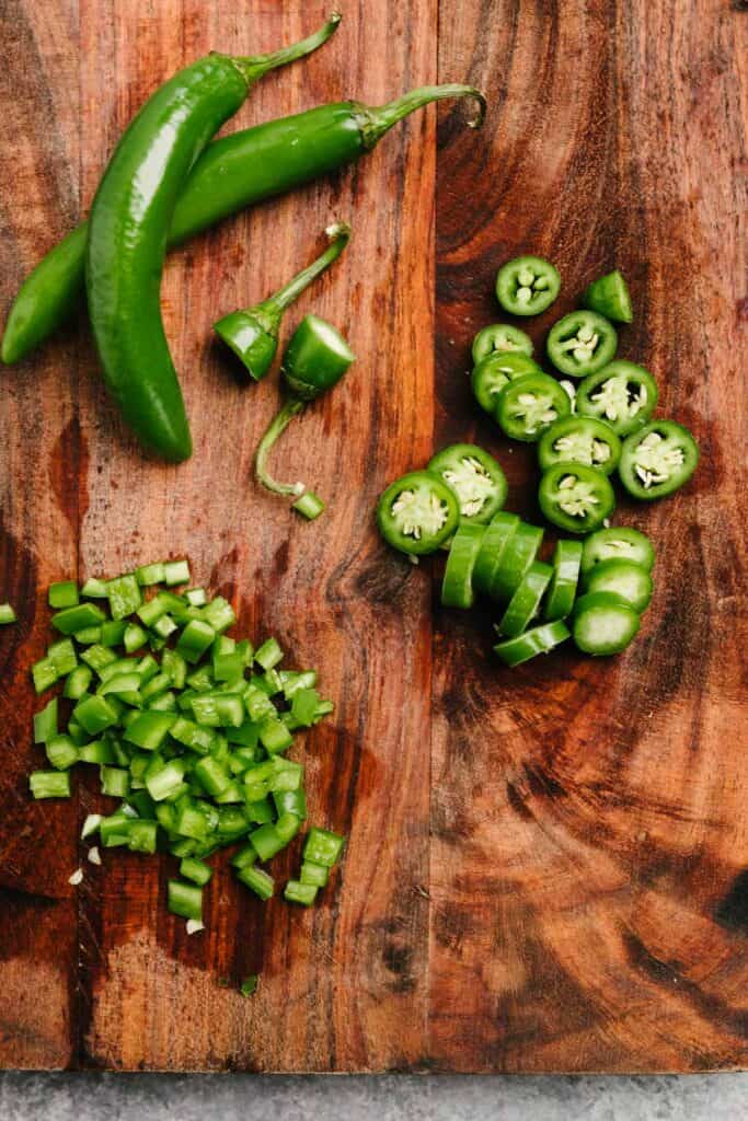 Two whole serrano chilies, one thinly sliced chile, and one seeded and minced chile on a wood cutting board.