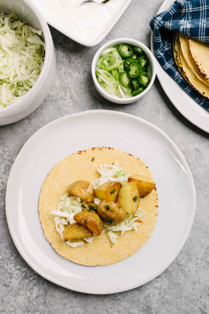 A corn tortilla layered with lime coleslaw, sour cream, and spicy potatoes on a white plate.