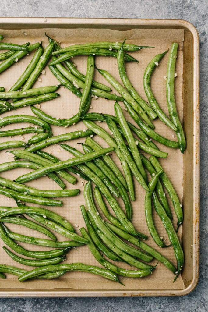 Green beans tossed with olive oil, garlic, salt and pepper on a parchment lined baking sheet.