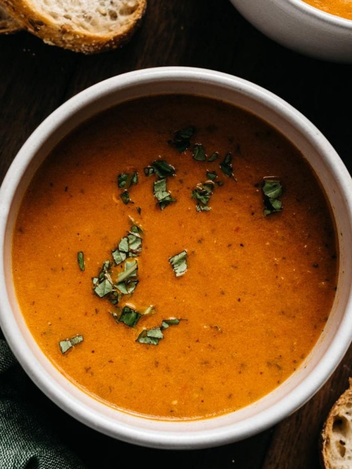 A bowl of roasted tomato soup garnished with fresh basil with slices of sourdough bread on the side.