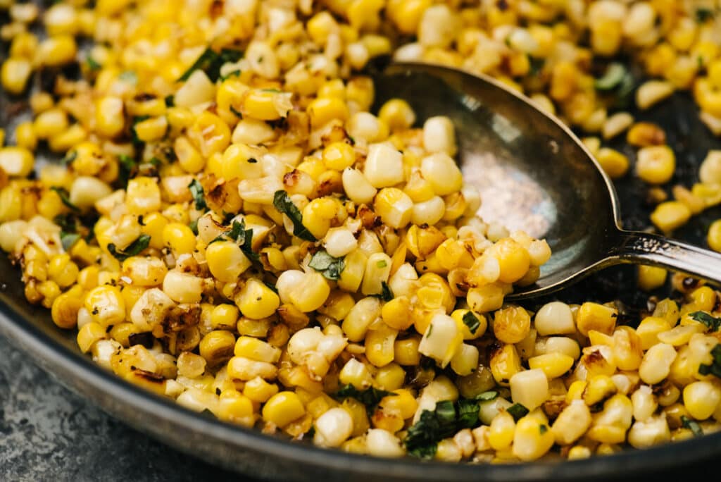 Side view, sauteed fresh corn in a skillet with fresh herbs and a silver spoon.