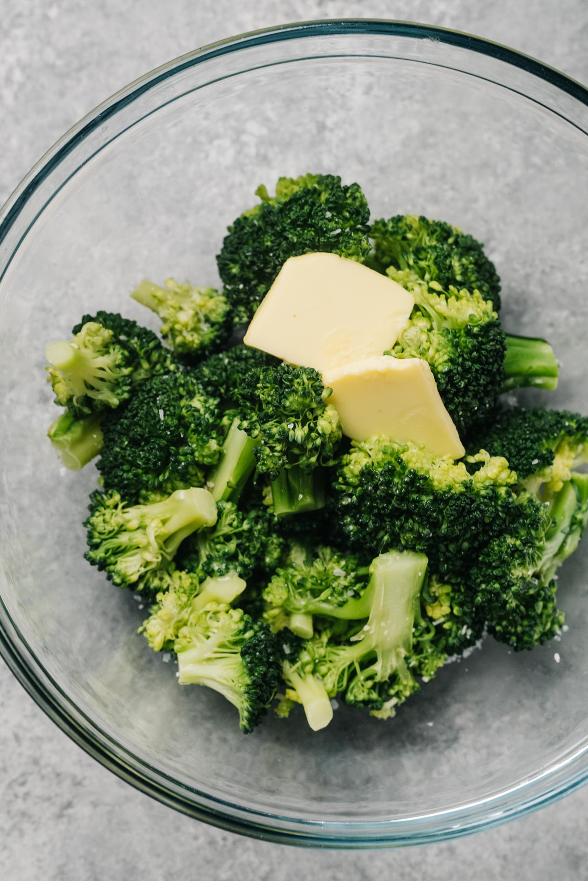 Two pats of butter melting over steamed broccoli in a glass bowl.