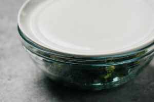 Broccoli florets in a glass microwave safe bowl with water and salt, covered with a plate for steaming.