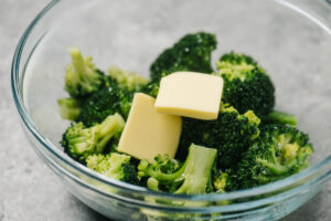 Steamed broccoli florets in a glass bowl with two pats of butter.