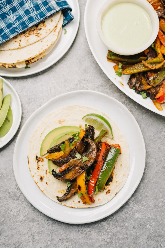 A corn tortilla on a white plate topped with vegetarian fajita filling, avocado slices, and limes.