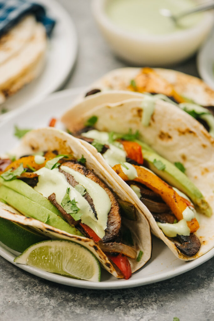 Side view, three vegetarian fajitas with cilantro sour cream on a white plate.